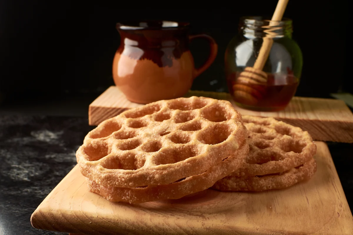 En una tabla de madera hay buñuelos de viento y a lo lejos se encuentra una taza con café de olla y un tarro con miel.