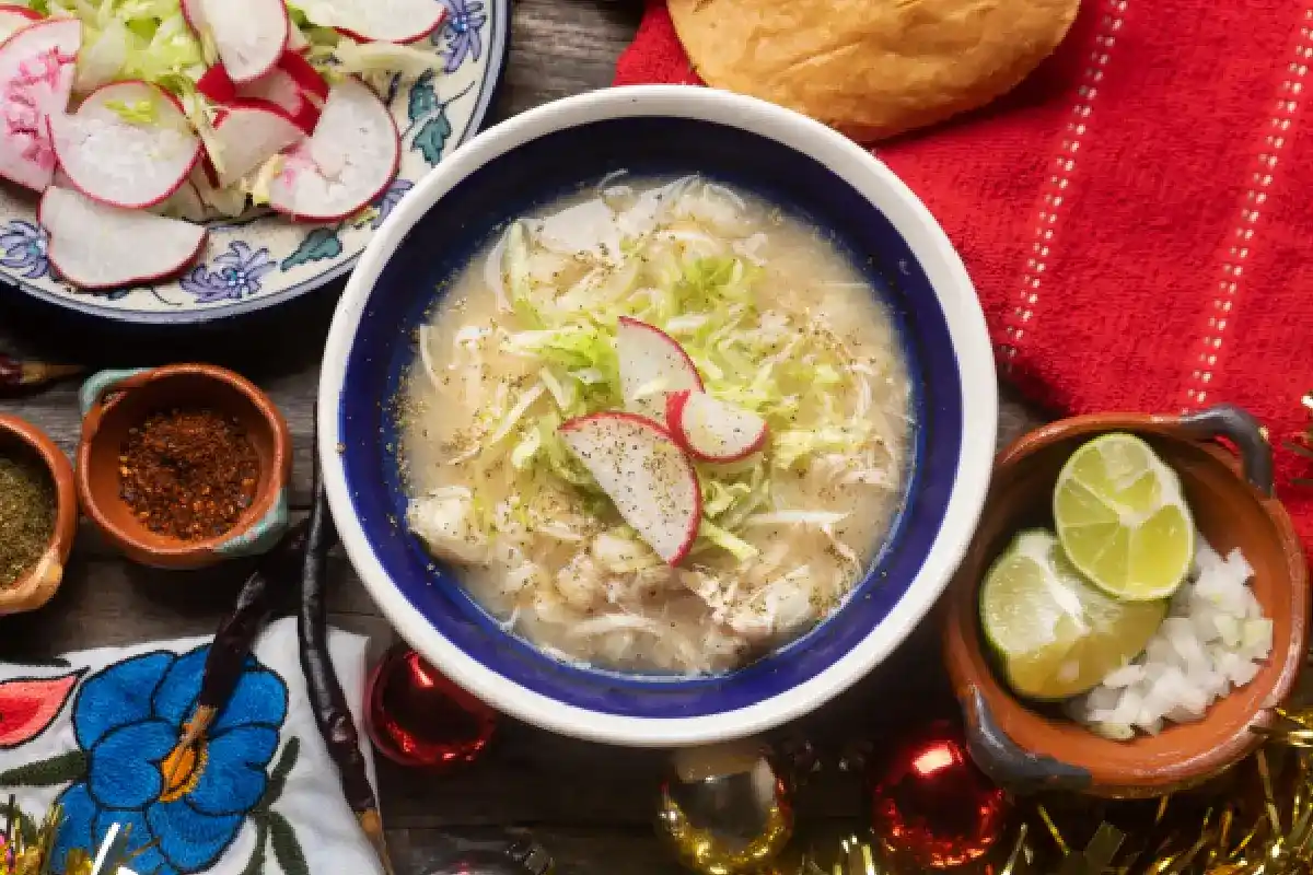 mesa con plato de pozole blanco preparado con cebolla, limón y lechuga