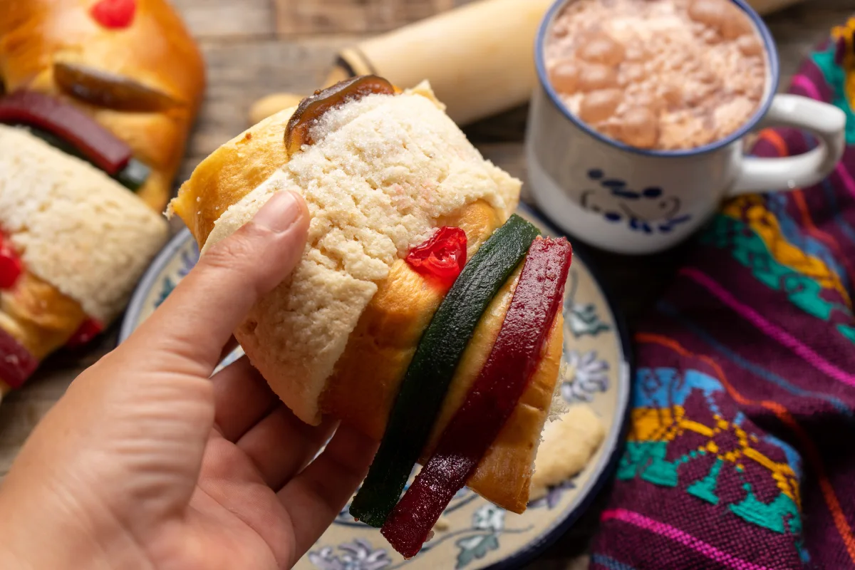 Una mano sostiene una rebanada de Rosca de Reyes y sobre la mesa hay una taza de chocolate caliente.