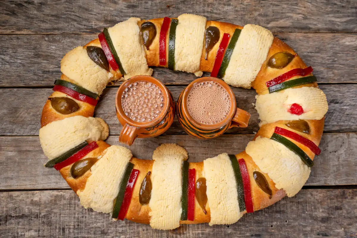 Sobre una mesa está una rosca de reyes completa y al centro hay dos tazas de chocolate caliente.