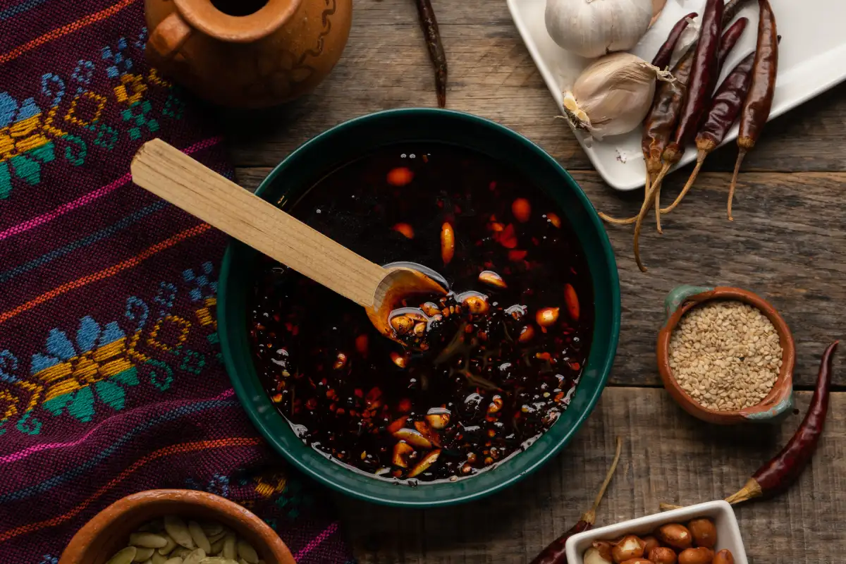 En un plato verde está servida abundante salsa macha y a su alrededor hay ingredientes como chile, pepita, ajonjoli y ajo.