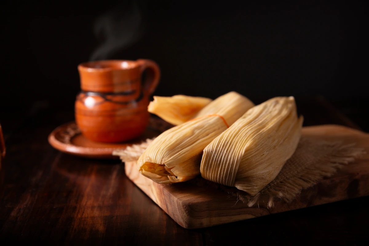 En una mesa están colocados dos tamales con hoja de maíz y una tarro con atole.