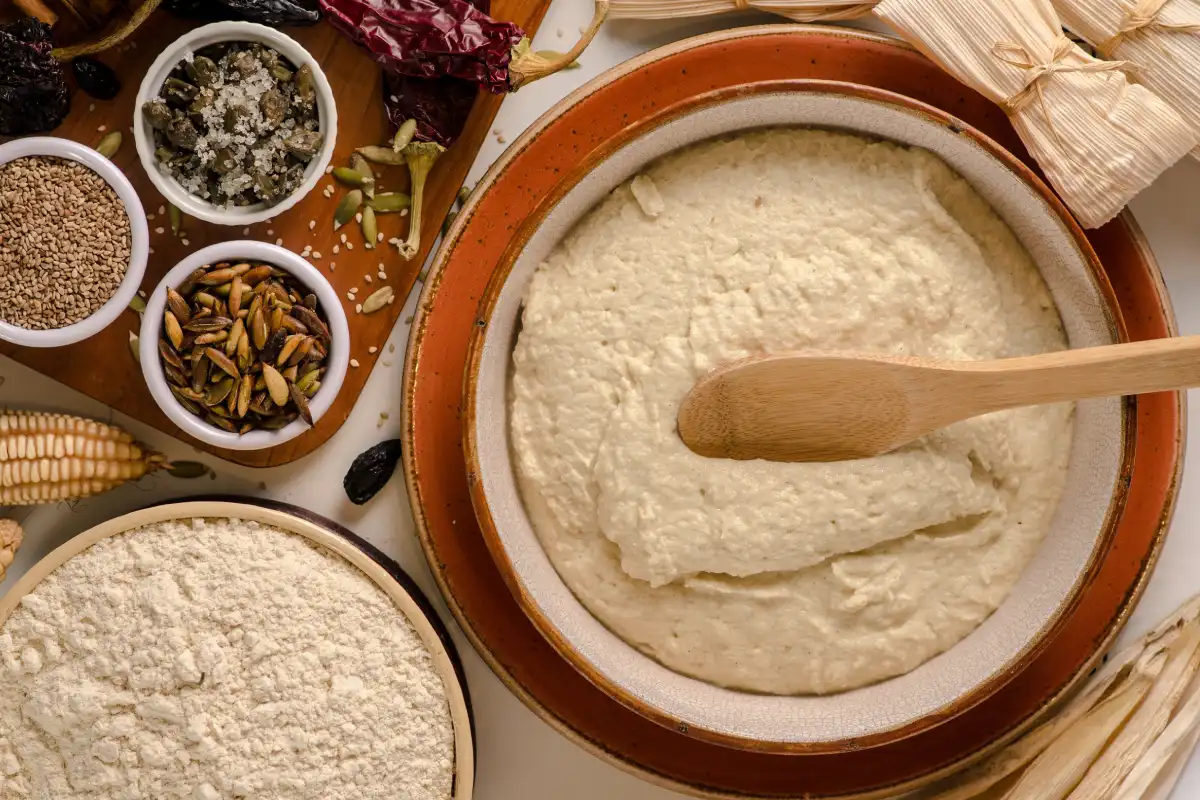 En un recipiente se encuentra servida la masa para preparar tamales, a un costado hay hojas secas de tamal y semillas.