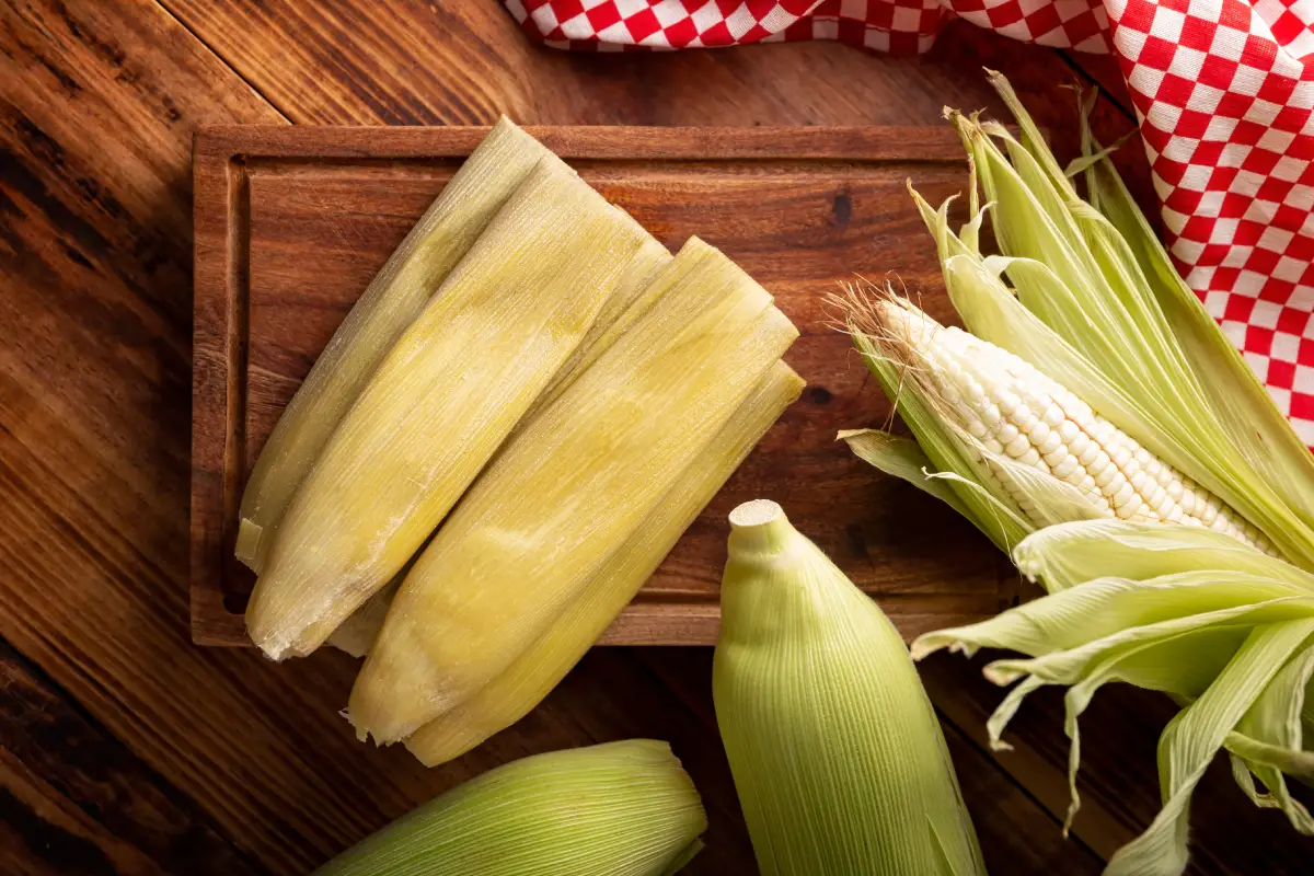 Sobre una mesa se encuentran tamales cerrados y a un costado hay mazorcas de elote.
