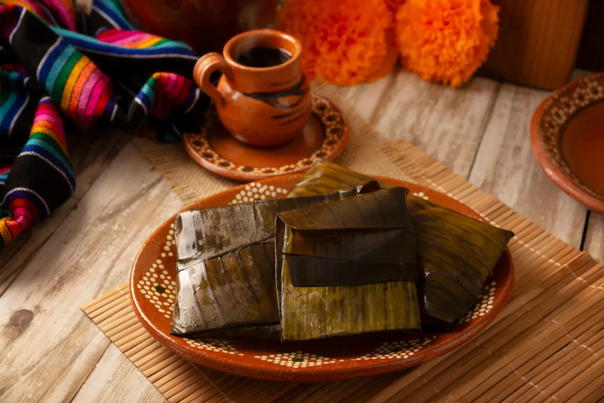Sobre un plato de barro están servidos tamales oaxaqueños con hojas de plátano y a un costado hay un jarrito de barro con café.