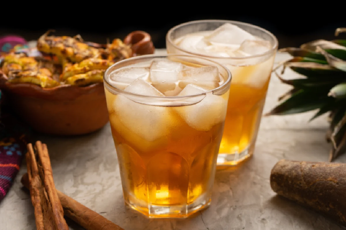 Sobre dos vasos con hielo está servido el tepache y a un costado se localiza la cáscara de piña, canela y piloncillo.