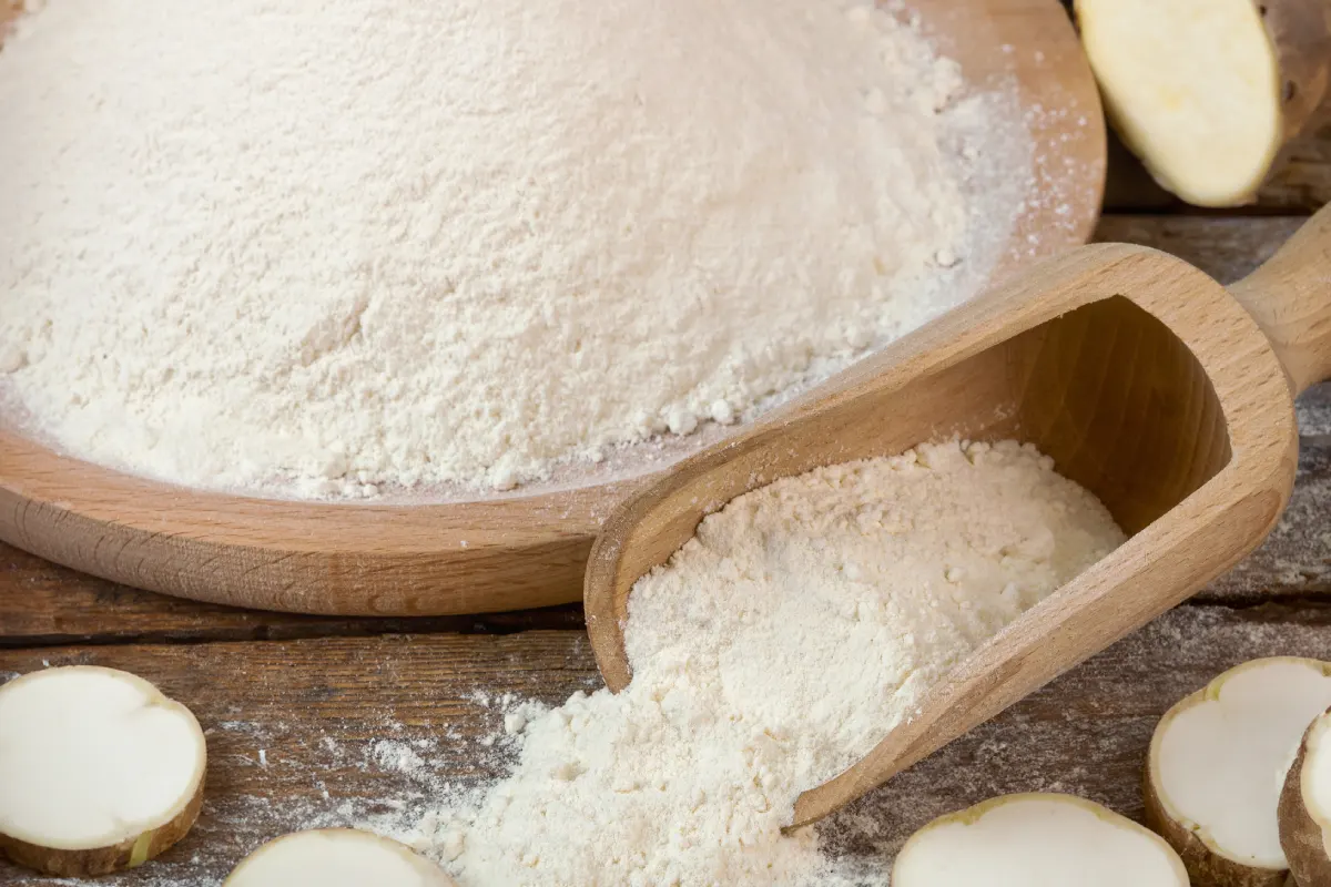 La harina de tapioca se derrama desde una cuchara de madera sobre una mesa con la raíz de tapioca en rodajas al fondo.