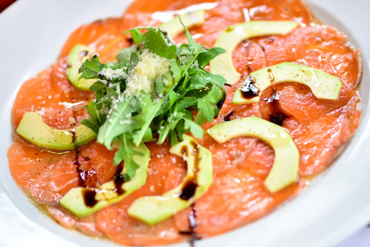 Está servido un carpaccio de salmón con aguacate en un plato blanco.