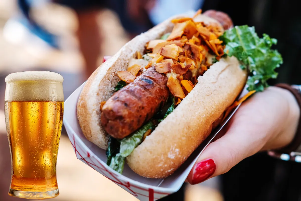 Una mano sostiene un hot dog hecho con salchicha vegana y a un costado está el recorte de una cerveza clara.