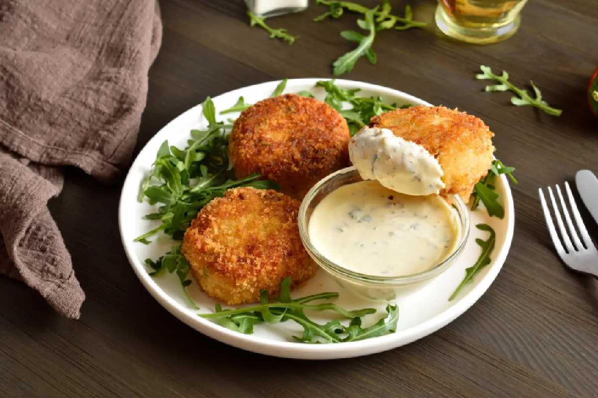 En un plato blanco están servidas tres croquetas de sardinas con mayonesa junto con un poco de arúgula.