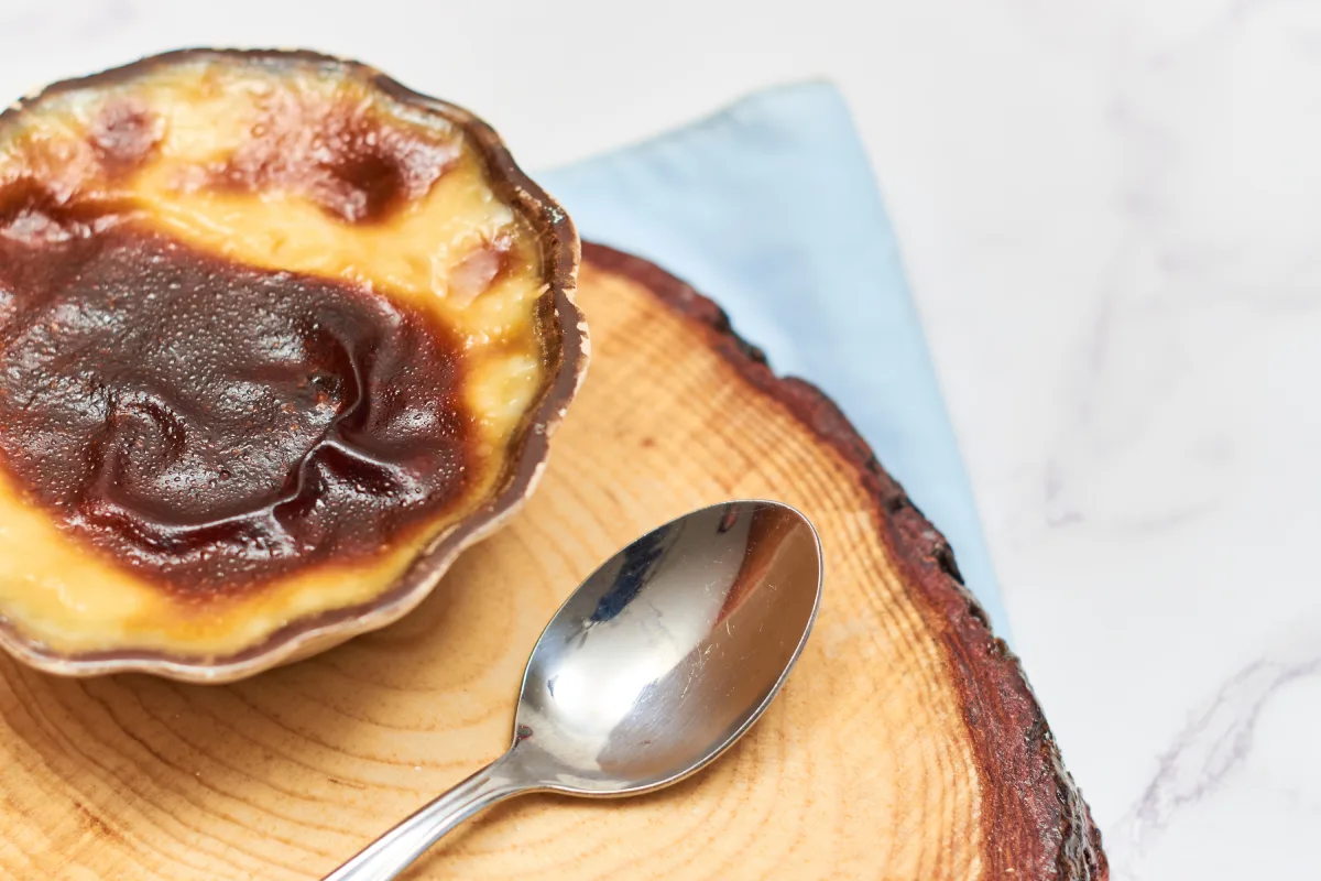 Sobre un pedazo de madera hay un recipiente con jericalla y una cuchara que lo acompaña.