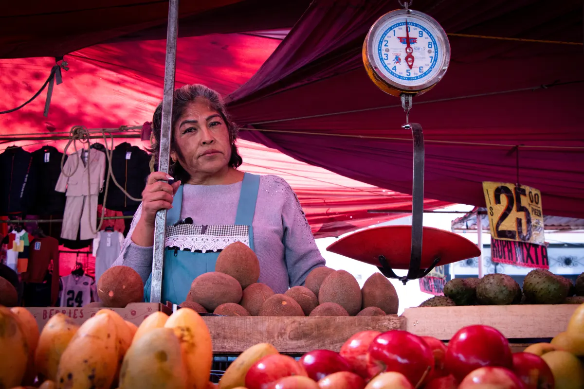 Comerciante se sostiene de un tubo a un costado está la báscula con la pesa la fruta que vende.